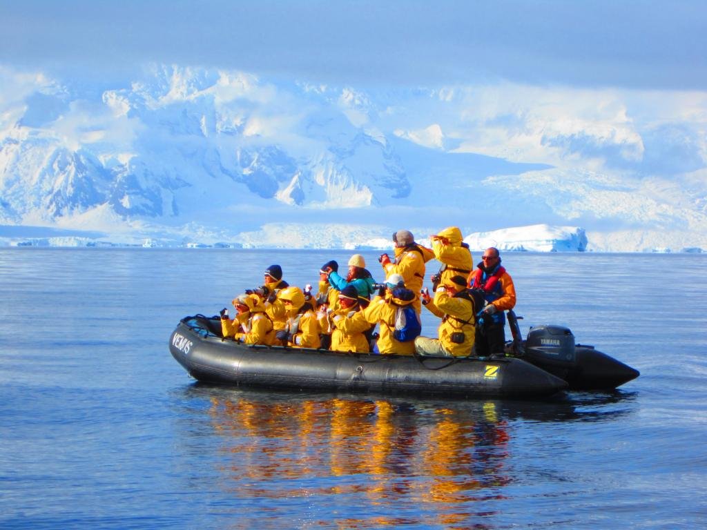 Zodiac cruise passengers in bright yellow parkas explore the Antarctic coastline, framed by towering glaciers and snow-covered peaks